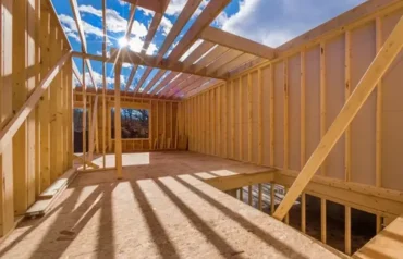 A construction worker wearing a hard hat and tool belt examines blueprints inside a wooden house frame, representing professional framing services.