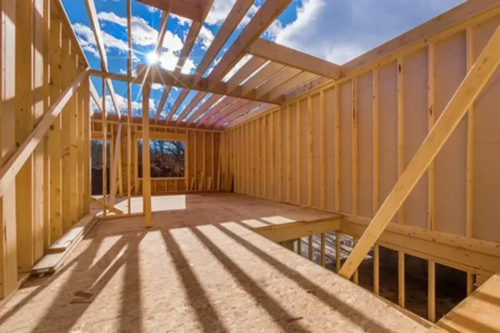 A construction worker wearing a hard hat and tool belt examines blueprints inside a wooden house frame, representing professional framing services.