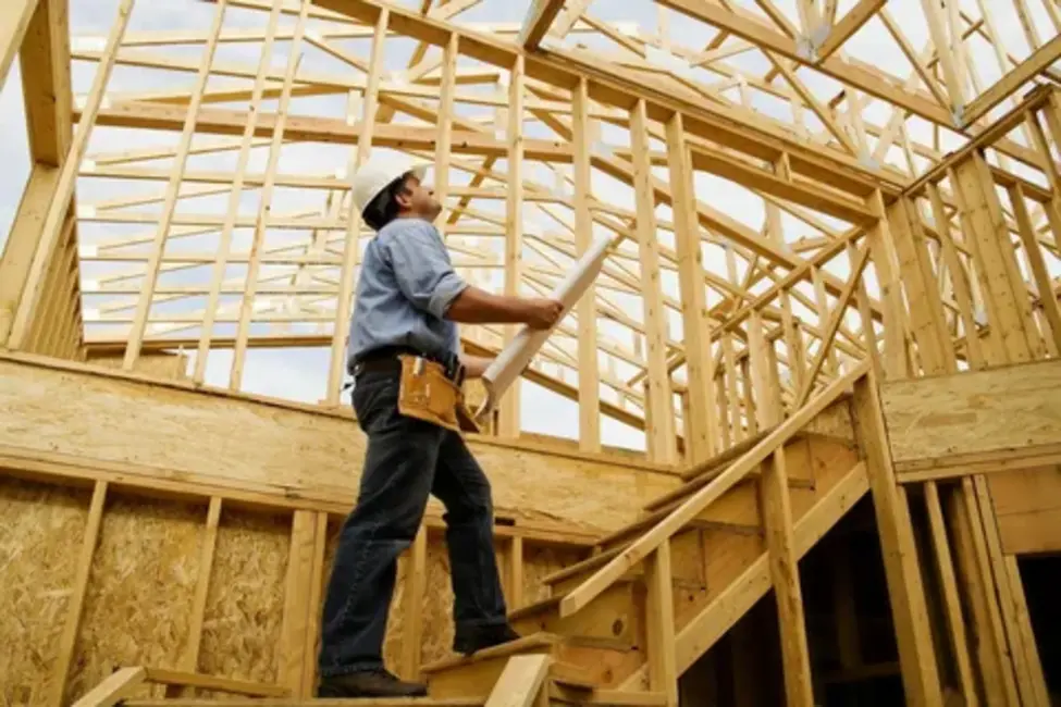  Construction worker holding blueprints while inspecting the wooden frame of a house under construction, walking up unfinished stairs.