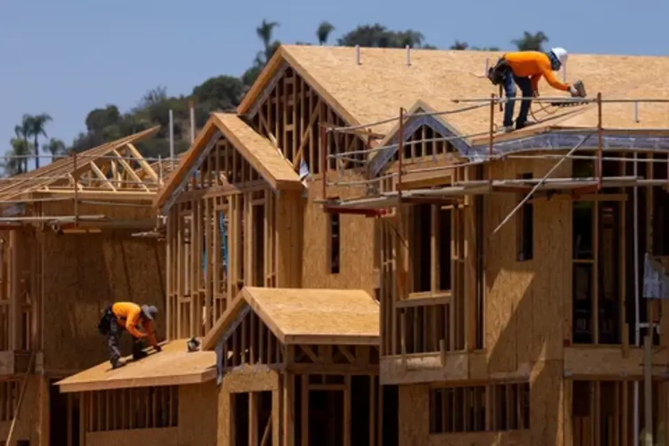 Construction workers in orange shirts working on the wooden frame and roof of a new residential building under development.