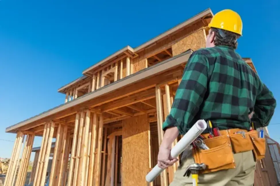 contractor with a hard hat and tool belt reviewing construction plans inside a partially built wooden house frame