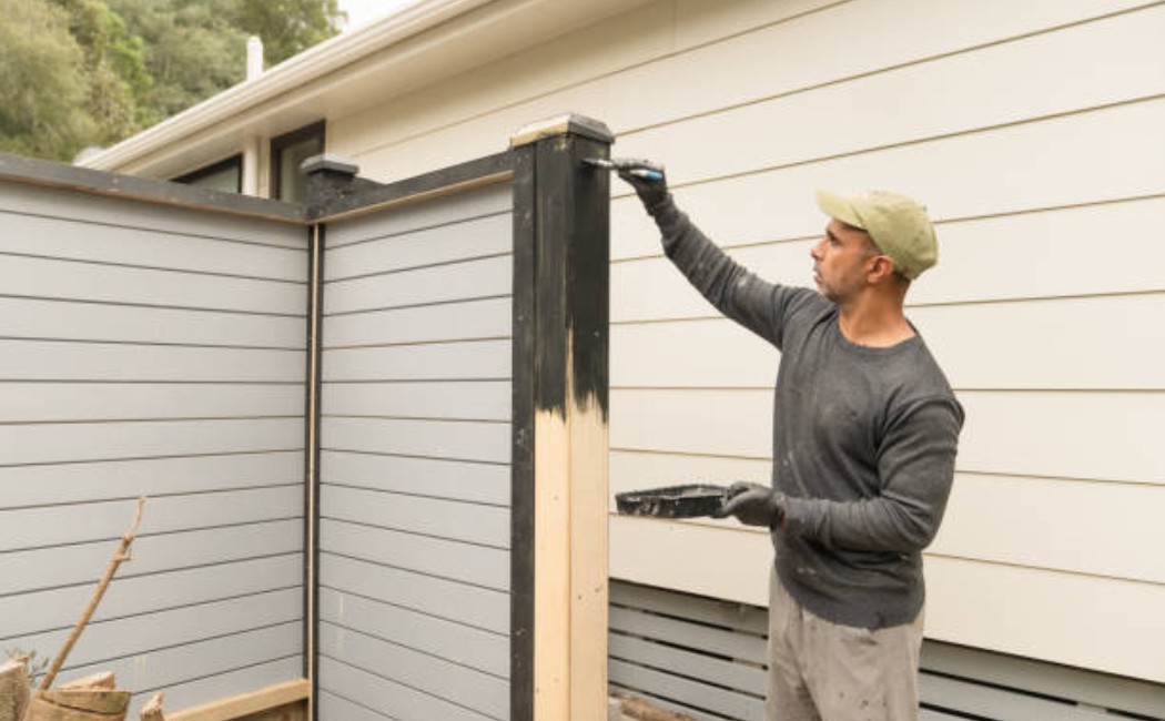Man applying wood stain to a wooden fence outdoors using a brush.